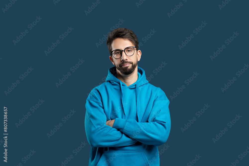 A young man with facial hair is standing confidently with his arms crossed, wearing a blue hoodie that matches the solid blue backdrop. His expression is serious and contemplative. A young man with facial hair is standing confidently with his arms crossed, wearing a blue hoodie that matches the solid blue backdrop. His expression is serious and contemplative.