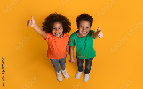 A cheerful African American boy and girl are standing on a bright yellow surface, looking upwards into the camera with joyful smiles, giving a thumbs up gesture, top view