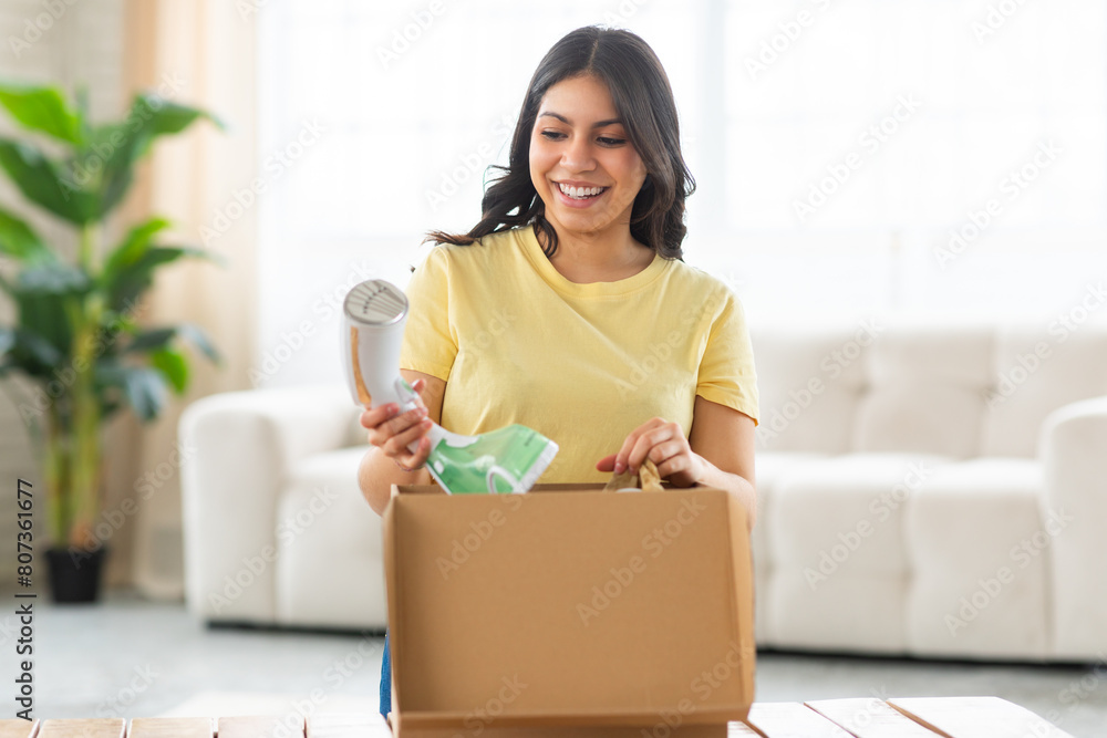 A cheerful middle eastern woman is standing in a brightly lit living room, looking delighted as she unpacks modern iron from a cardboard box.