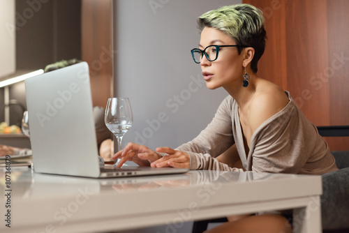 a beautiful girl with short hair and glasses is sitting indoors at a laptop chatting and working online