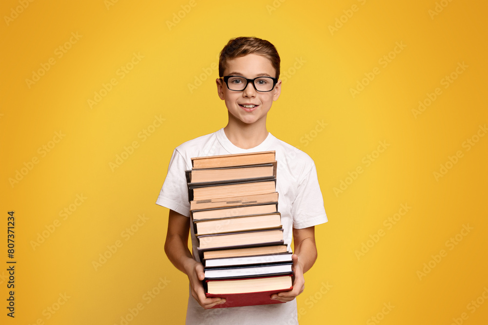 Cute diligent student boy in glasses with heap of books in hands, orange studio background Cute diligent student boy in glasses with heap of books in hands, orange studio background