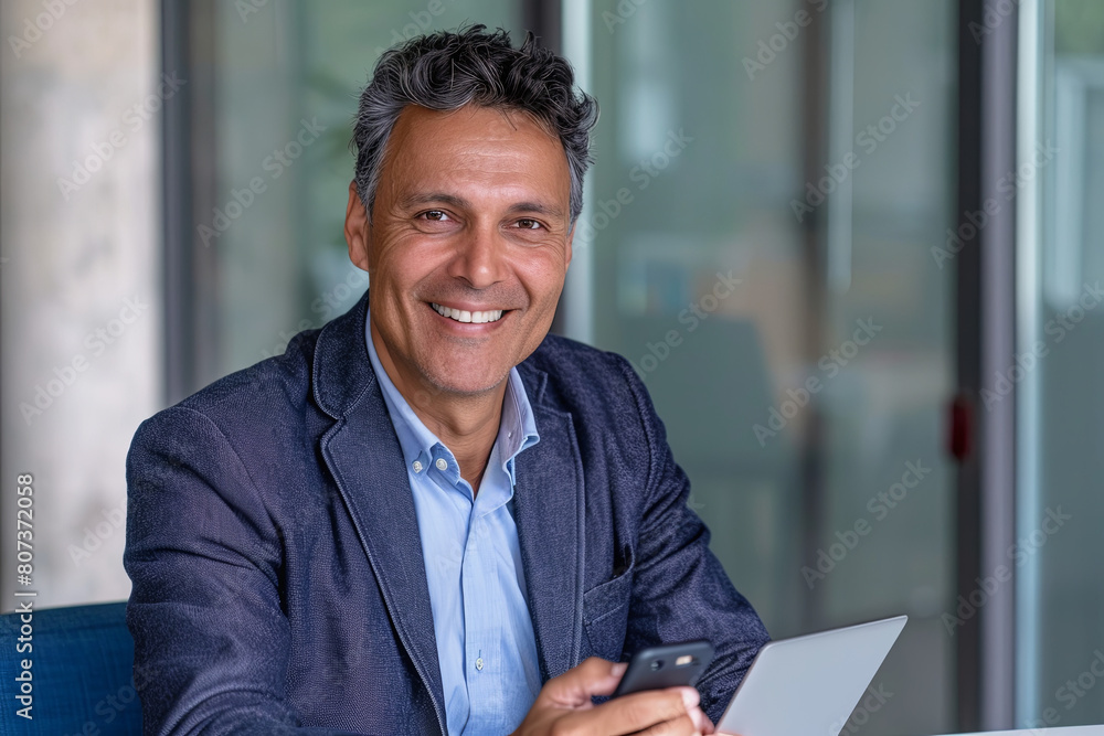 Fototapeta premium Middle aged Hispanic business manager ceo using cell phone mobile app, laptop. Smiling Latin or Indian mature man businessman holding smartphone 