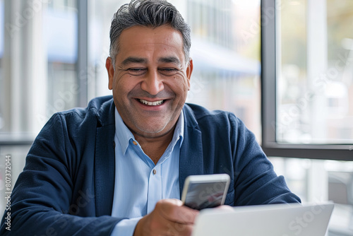 Middle aged Hispanic business manager ceo using cell phone mobile app, laptop. Smiling Latin or Indian mature man businessman holding smartphone
