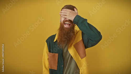 A cheerful, young, redhead man with a beard, laughing while covering his eyes, stands against an isolated yellow background. © Krakenimages.com
