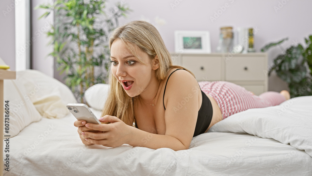 A surprised blonde woman lies on a bed with a phone in a cozy bedroom. A surprised blonde woman lies on a bed with a phone in a cozy bedroom.