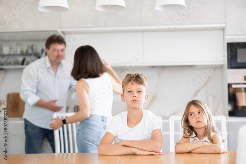 Brother and sister sitting at table in kitchen during conflict between their parents