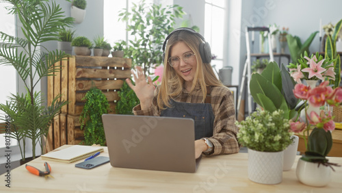 A smiling woman with headphones videoconferencing in a flower shop surrounded by plants and a laptop.