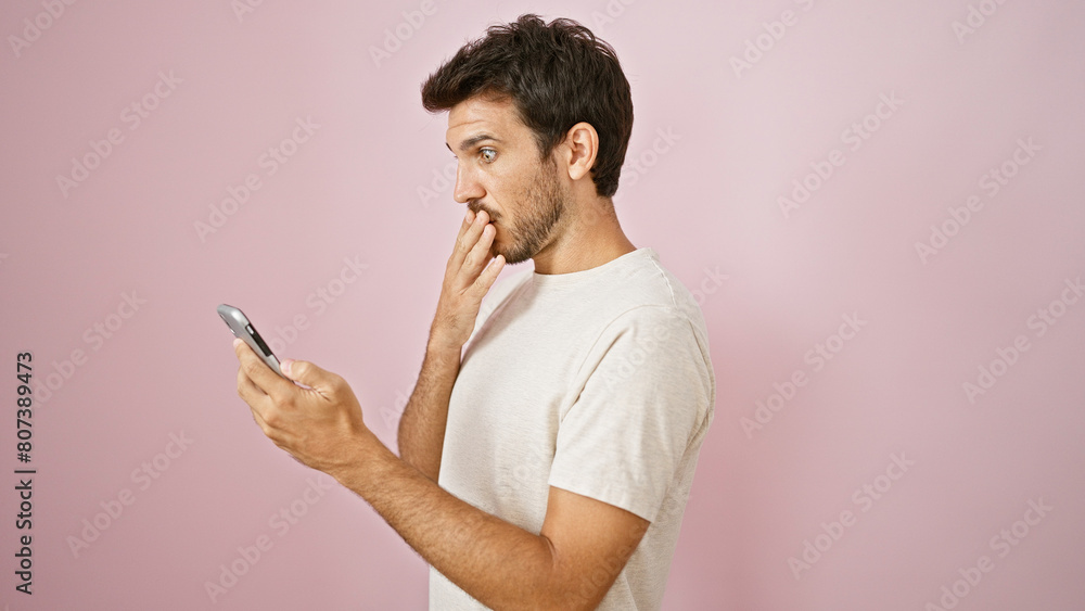 A surprised hispanic man with a beard looks at his phone against a pink background. A surprised hispanic man with a beard looks at his phone against a pink background.