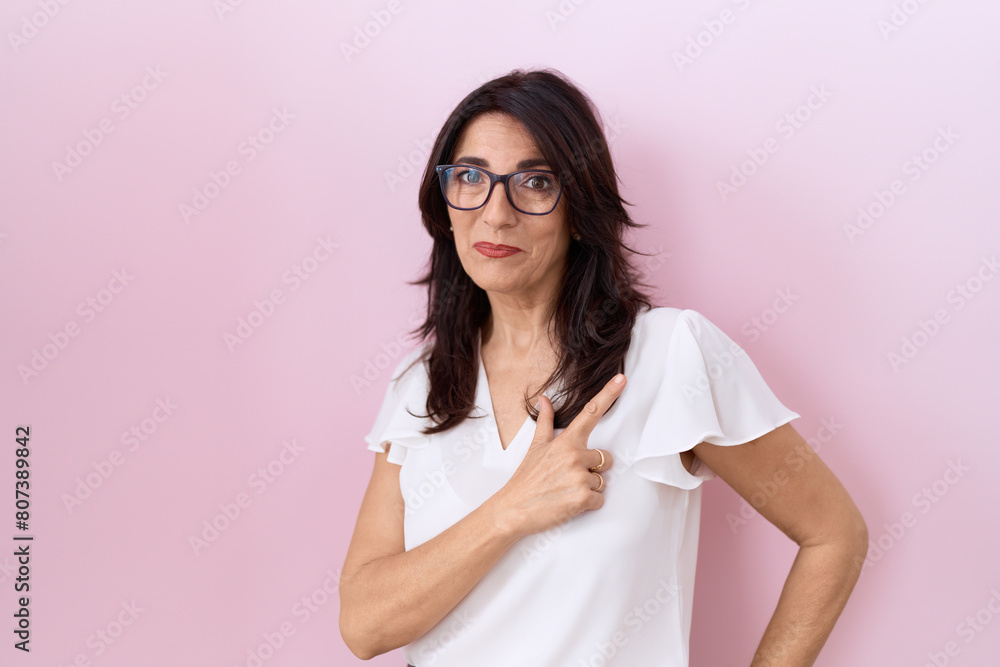 Middle age hispanic woman wearing casual white t shirt and glasses cheerful with a smile on face pointing with hand and finger up to the side with happy and natural expression Middle age hispanic woman wearing casual white t shirt and glasses cheerful with a smile on face pointing with hand and finger up to the side with happy and natural expression