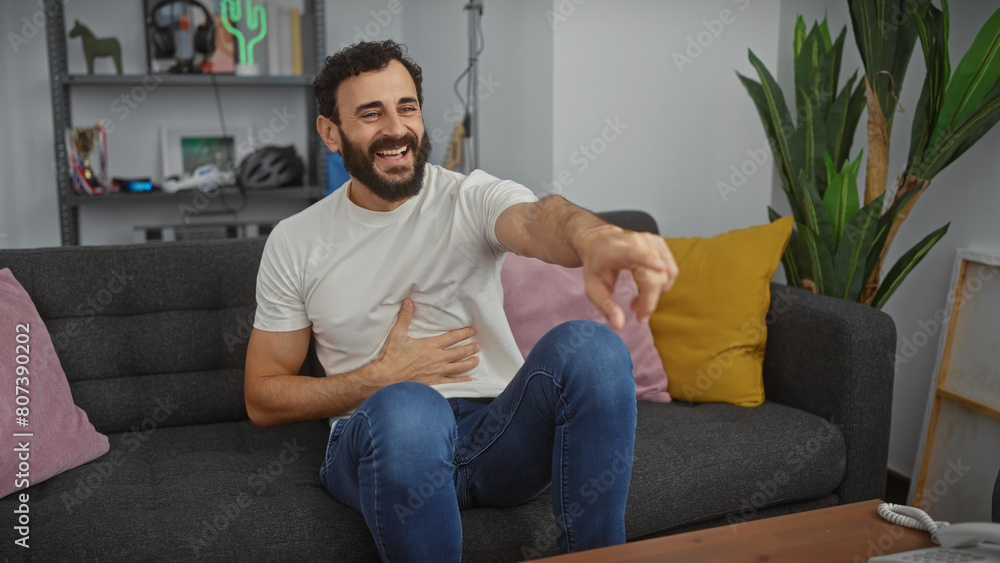 Laughing man with beard sitting on sofa in modern living room pointing at camera Laughing man with beard sitting on sofa in modern living room pointing at camera