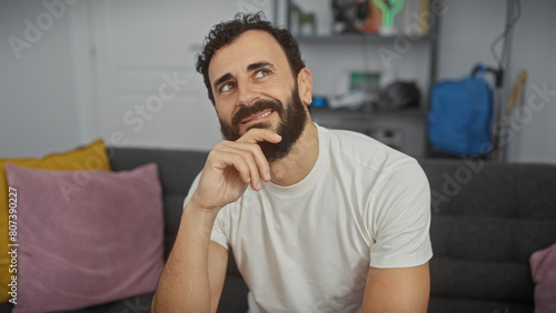 A pensive middle-aged man with a beard sits in a modern living room, reflecting with a smile. photo