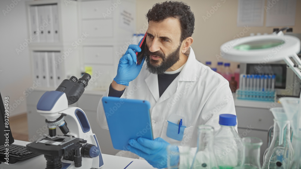 A mature bearded man in a lab coat talks on a phone and uses a tablet in a science laboratory, showing multitasking and technology use. A mature bearded man in a lab coat talks on a phone and uses a tablet in a science laboratory, showing multitasking and technology use.