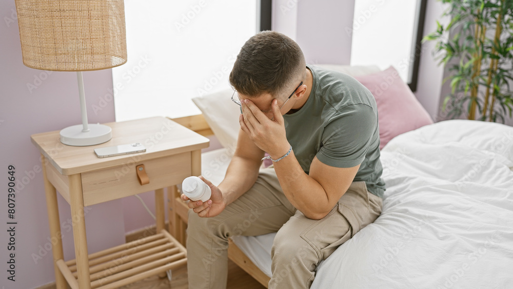 A distressed hispanic man sits on a bed holding a medicine bottle in a modern bedroom. A distressed hispanic man sits on a bed holding a medicine bottle in a modern bedroom.
