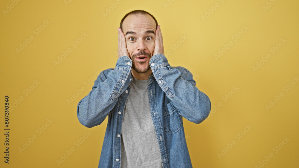 Obraz premium Surprised bald hispanic man with beard wearing denim expressing shock against a yellow isolated background.
