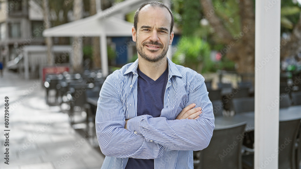 Naklejka premium A bearded, bald hispanic man crosses his arms while standing on a city street with an outdoor cafe in the background