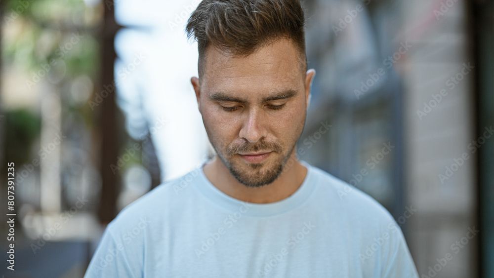 Close-up of a pensive young hispanic man with a beard, standing on a city street with a blurred urban background. Close-up of a pensive young hispanic man with a beard, standing on a city street with a blurred urban background.