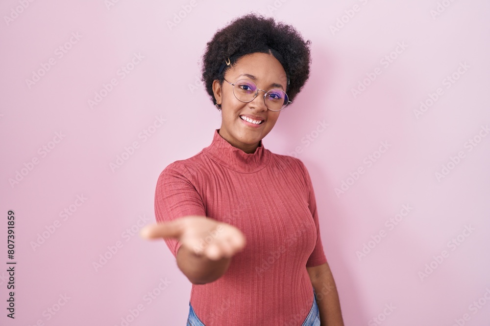 Beautiful african woman with curly hair standing over pink background smiling cheerful offering palm hand giving assistance and acceptance. Beautiful african woman with curly hair standing over pink background smiling cheerful offering palm hand giving assistance and acceptance.