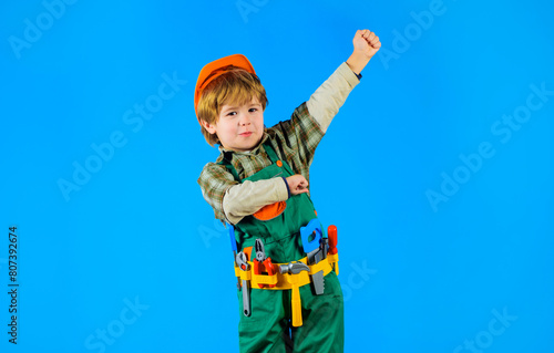 Little builder in construction helmet and tool belt. Kid repairman in hard hat and overalls with tool belt of toy tools for building. Smiling child boy in uniform with set of toy construction tools.