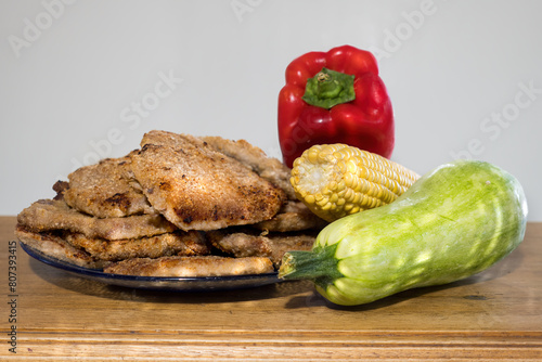 stack of breaded milanesas on a plate placed on a wooden table next to a corn, a red pepper and a zuchini squash.