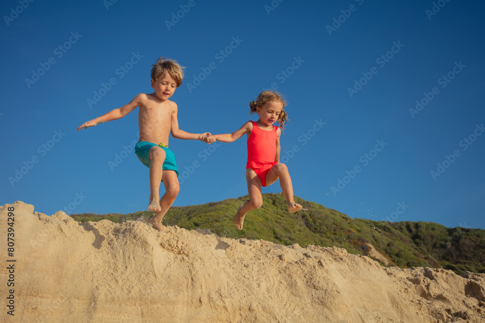 Two cheerful kids are engaging in playful jump from sandy dune Two cheerful kids are engaging in playful jump from sandy dune