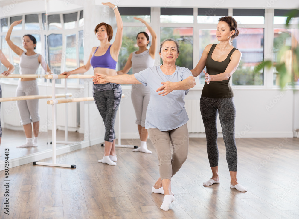 Focused female ballet choreographer patiently instructing senior woman in basic ballet techniques at group dance class for amateurs in spacious light-filled studio..
