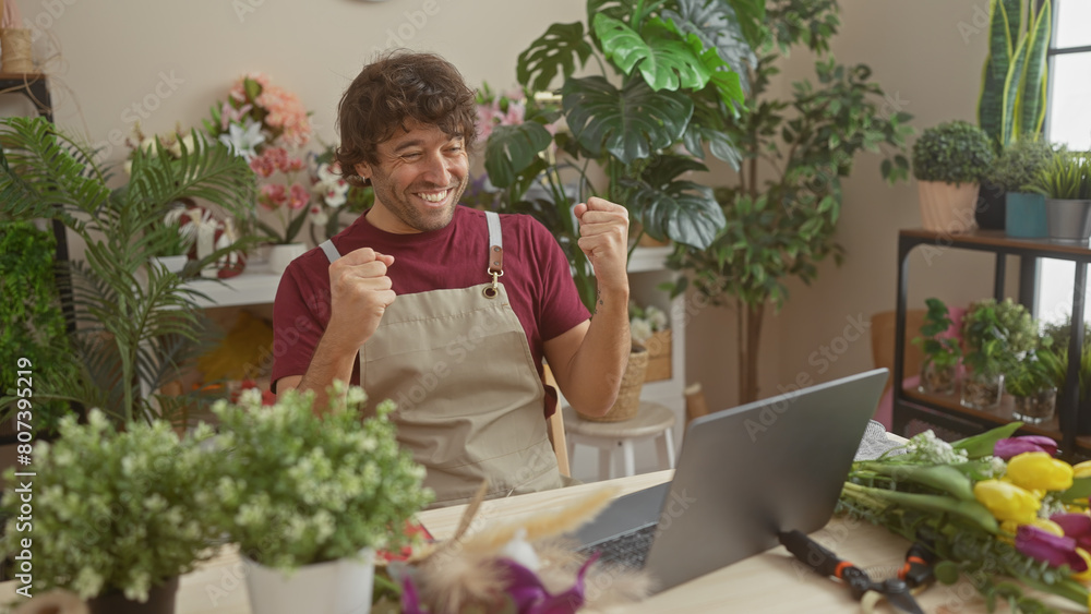 A smiling young hispanic man in a florist shop with a laptop, surrounded by green plants and colorful flowers.