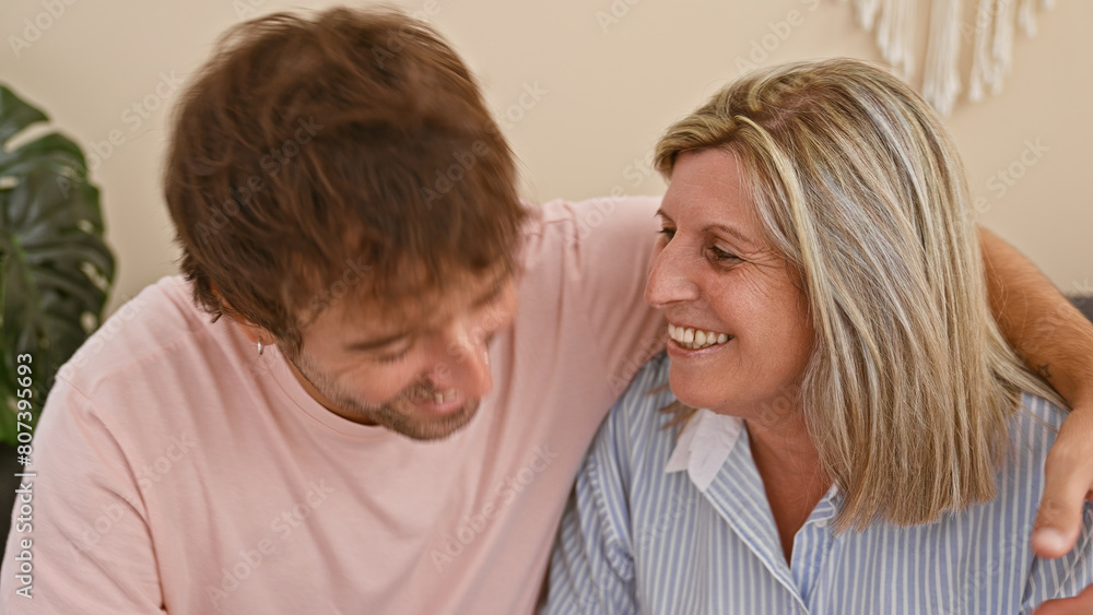 Heartwarming mother and son sitting together on the living room sofa, joyfully hugging and smiling in their cozy home, radiating happiness and confidence in their casual lifestyle. Heartwarming mother and son sitting together on the living room sofa, joyfully hugging and smiling in their cozy home, radiating happiness and confidence in their casual lifestyle.