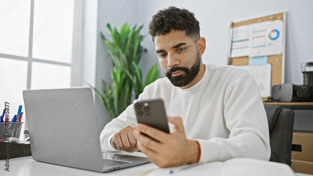 Hispanic man with beard using smartphone at office desk with laptop and indoor plant. Hispanic man with beard using smartphone at office desk with laptop and indoor plant.