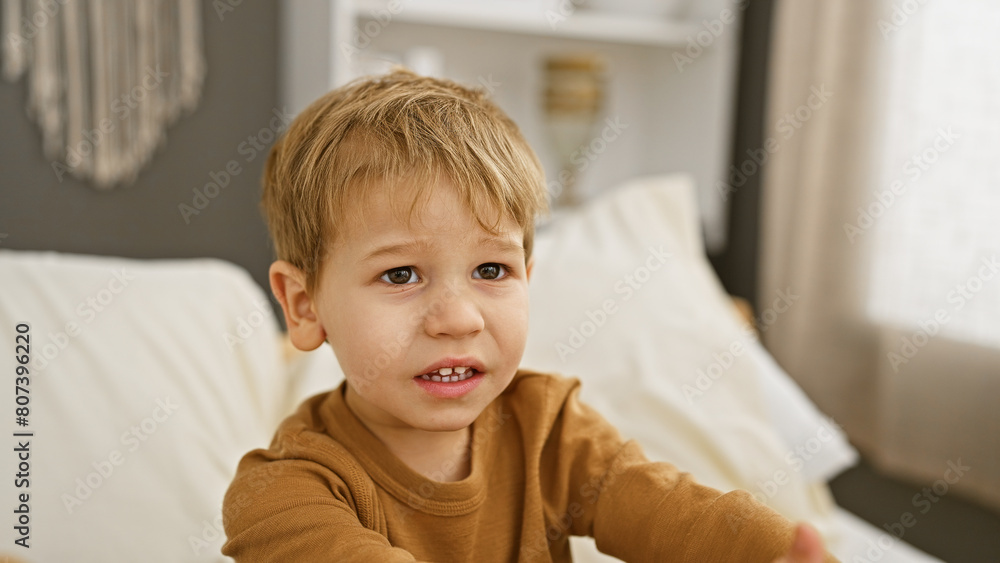 Upset blond toddler boy sitting on a white bed in a home bedroom interior Upset blond toddler boy sitting on a white bed in a home bedroom interior