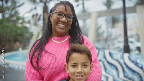 A smiling woman in a pink sweater embracing a young boy outdoors with palm trees and a pool in the background, depicting a warm family moment. photo