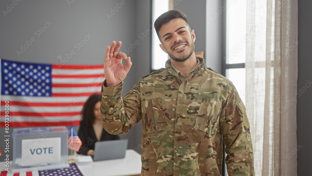 A smiling soldier in camo making an 'ok' gesture indoors, with a woman and us flag in the background at a voting center A smiling soldier in camo making an 'ok' gesture indoors, with a woman and us flag in the background at a voting center