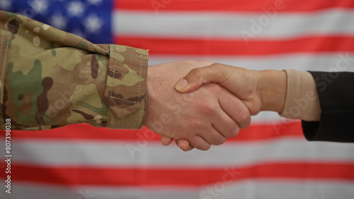 A civilian woman and a military man shake hands in front of an american flag, symbolizing collaboration.