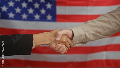 Man and woman shaking hands in an office with an american flag backdrop, symbolizing cooperation.