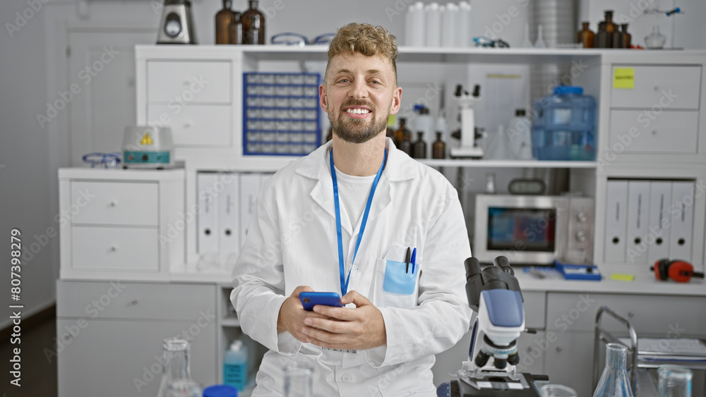A smiling young caucasian man with a beard and blue eyes wearing a lab coat using a smartphone in a laboratory. A smiling young caucasian man with a beard and blue eyes wearing a lab coat using a smartphone in a laboratory.