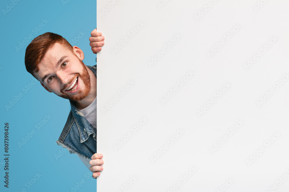 Cheerful young smiling guy looking out of white blank placard ready for text, information or product, blue background Cheerful young smiling guy looking out of white blank placard ready for text, information or product, blue background