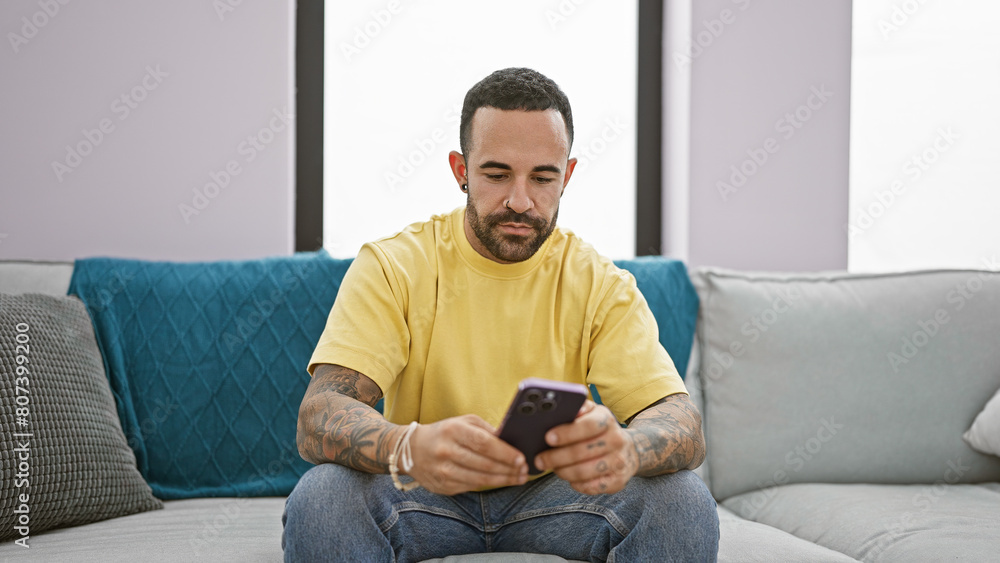 A focused young hispanic man with a beard sits at home using his smartphone, portraying a casual indoor lifestyle scene. A focused young hispanic man with a beard sits at home using his smartphone, portraying a casual indoor lifestyle scene.