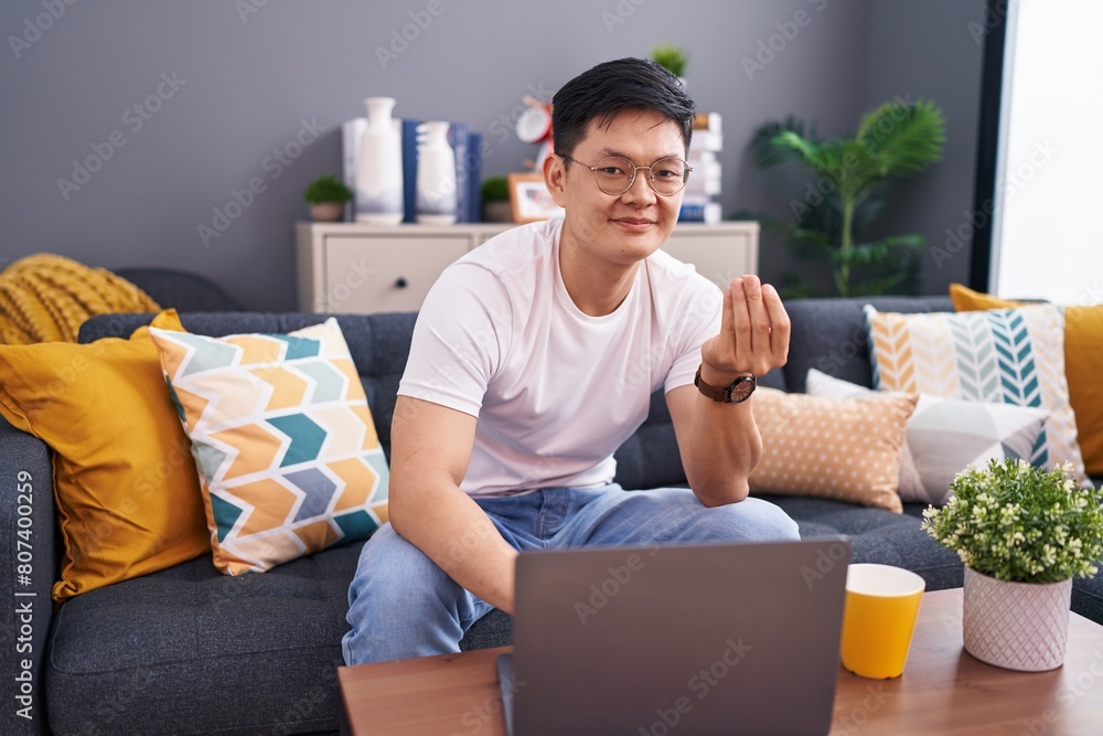 Young asian man using laptop at home sitting on the sofa doing money gesture with hands, asking for salary payment, millionaire business Young asian man using laptop at home sitting on the sofa doing money gesture with hands, asking for salary payment, millionaire business