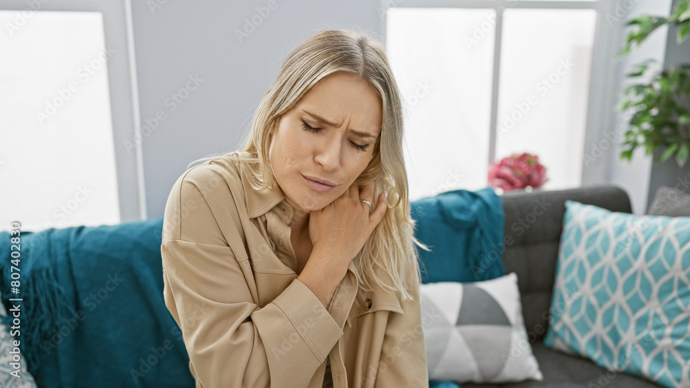 Worried and unhappy, young blonde woman suffers severe cervical pain sitting alone on sofa at home Worried and unhappy, young blonde woman suffers severe cervical pain sitting alone on sofa at home