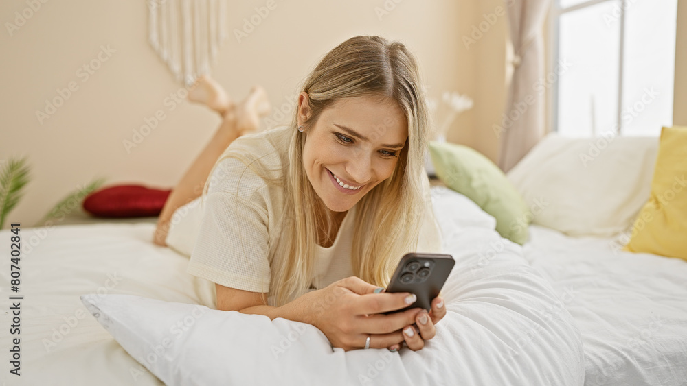 Confident young blonde woman enjoying morning in bed, smilingly using smartphone in a comfortable bedroom setting Confident young blonde woman enjoying morning in bed, smilingly using smartphone in a comfortable bedroom setting