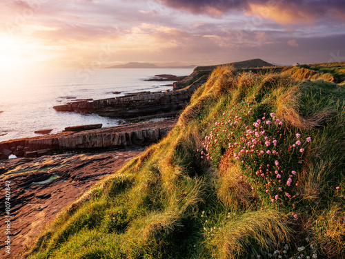 Stunning sunset nature scene with rough stone coastline, green grass hill, dramatic sky and mountains in the background. Mullaghmore head area, Ireland. Sun flare. Irish landscape. photo