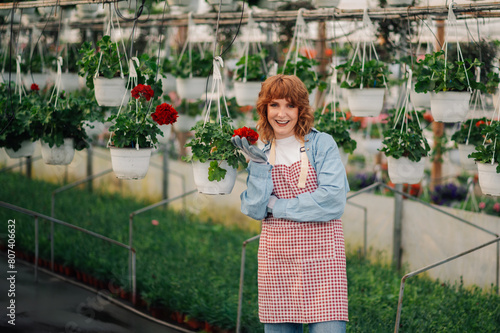 Young happy florist posing with potted hanging flowers at hothouse.