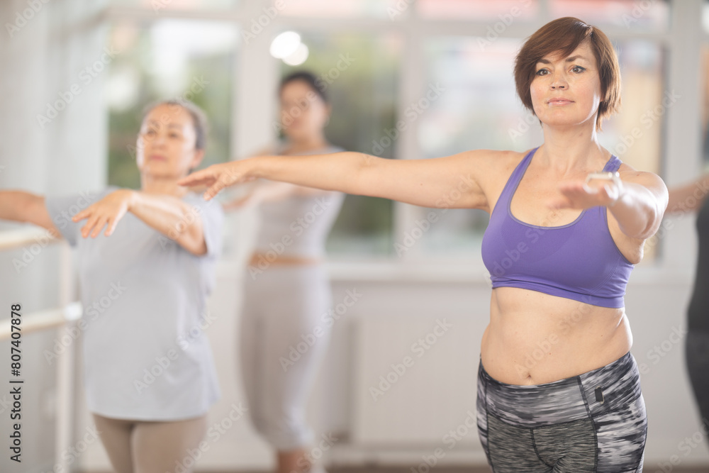 Positive middle aged woman, participating in beginner ballet class, practicing basic movements with female group of various ages in bright choreography studio with natural lighting