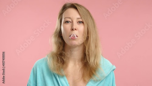 A playful young woman in a casual blue robe blows a large bubble with bubble gum, set against a soft pink background, showcasing a fun, carefree moment. 