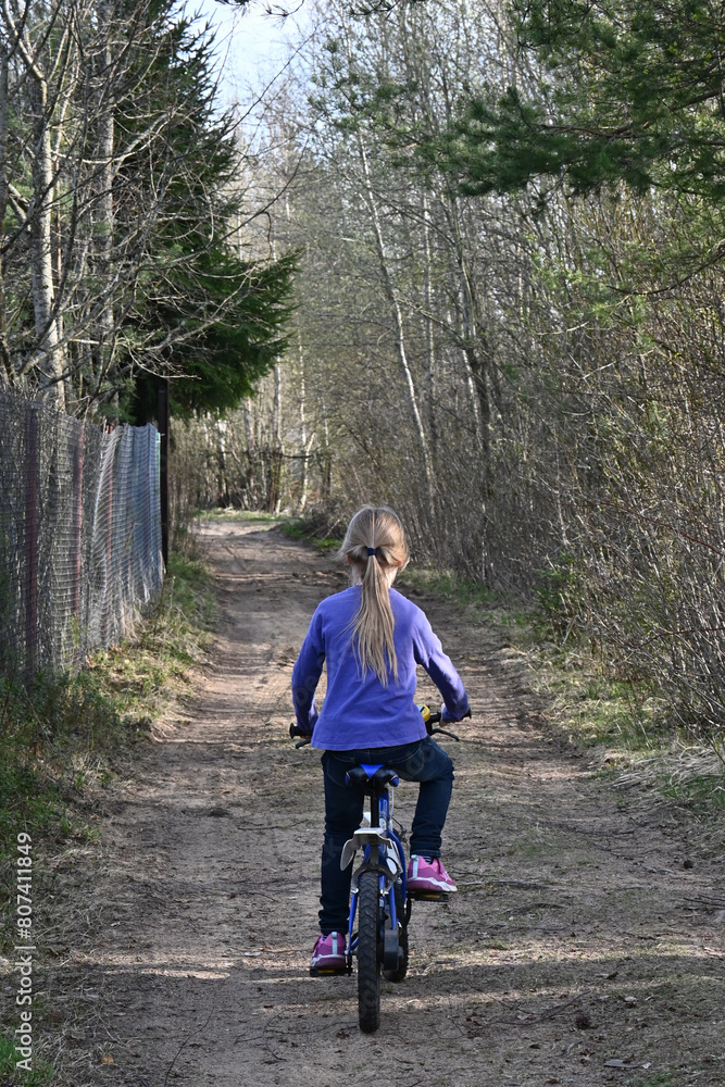 Fototapeta premium A little girl of 7 years old rides a blue bicycle on the road. Countryside, rustic landscape.