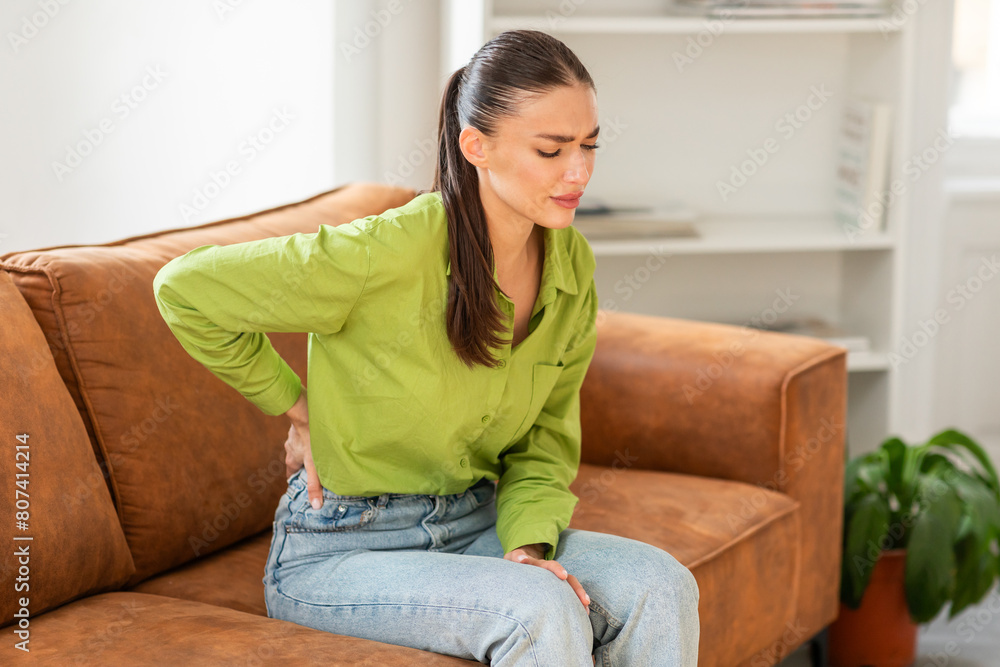 Young woman, dressed in green blouse and blue jeans, sits on a brown leather sofa with a pained expression, clutching her lower back with one hand, indicating discomfort or a potential back problem. Young woman, dressed in green blouse and blue jeans, sits on a brown leather sofa with a pained expression, clutching her lower back with one hand, indicating discomfort or a potential back problem.