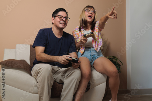 Latin adult couple playing video games on the couch in the living room, couple pastime, concept pointing her finger at the TV on beige background