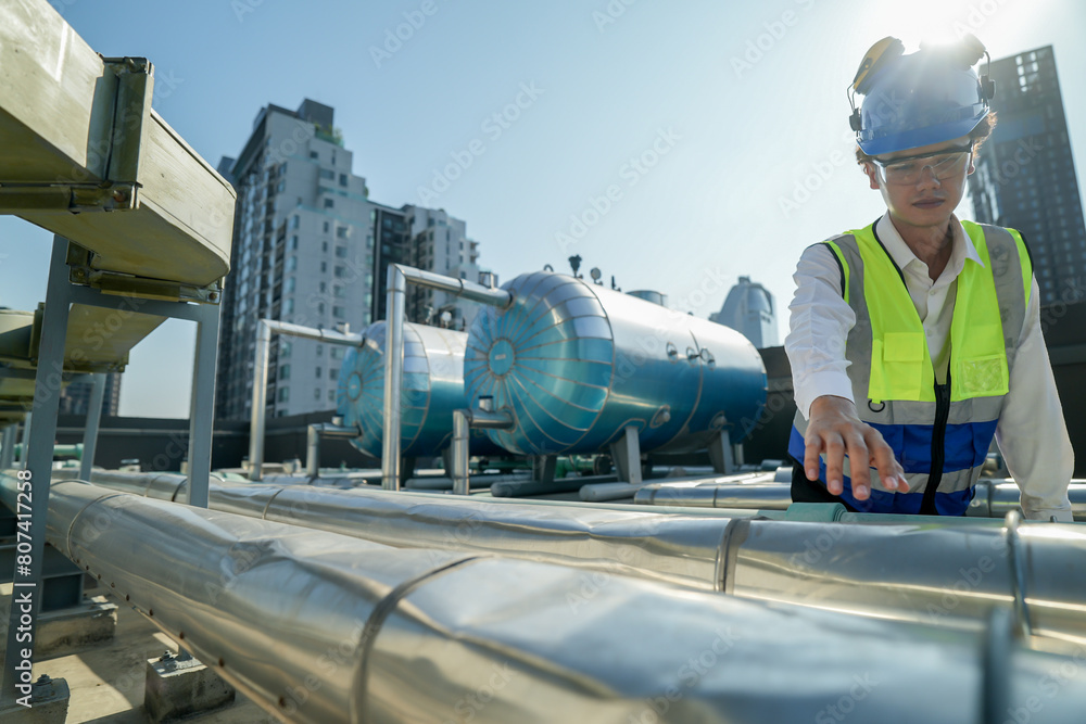 Serious engineer inspecting HVAC equipment with a tablet on a commercial building rooftop. A focused engineer wearing a safety helmet and vest inspects machinery on a city building’s rooftop.