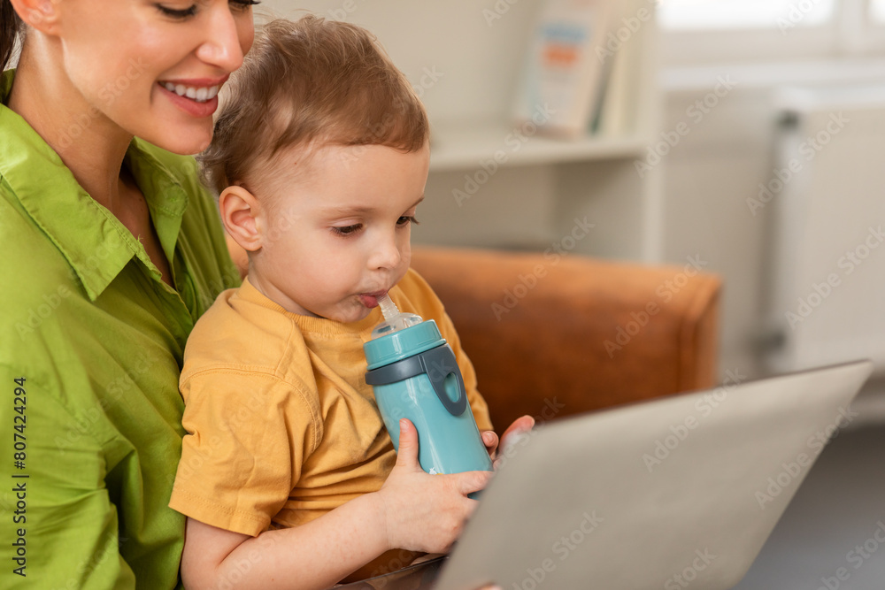 A woman is sitting while holding a child in her arms, multitasking by using a laptop. Mother and son watching cartoon together, home interior, copy space A woman is sitting while holding a child in her arms, multitasking by using a laptop. Mother and son watching cartoon together, home interior, copy space