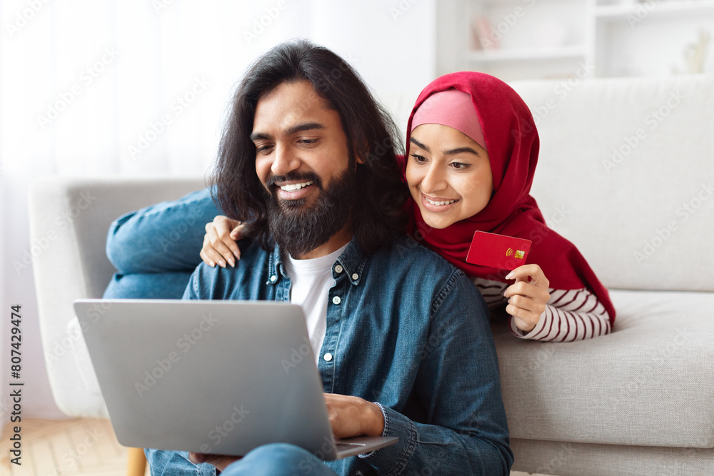 A young Muslim couple is comfortably seated on sofa in a well-lit living room, engaged in online shopping, with the man holding a laptop and the woman, wearing a red hijab, displaying a credit card A young Muslim couple is comfortably seated on sofa in a well-lit living room, engaged in online shopping, with the man holding a laptop and the woman, wearing a red hijab, displaying a credit card