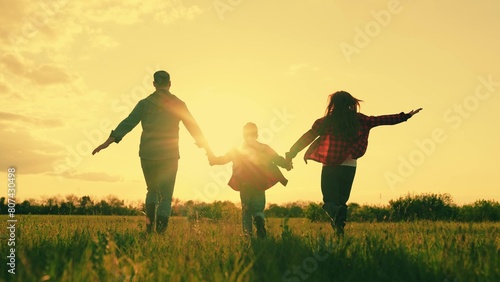 Happy family running flying holding hands at sunset sunny field back view slow motion. Mother father and son playing movement on natural grass meadow enjoy freedom together lifestyle leisure activity